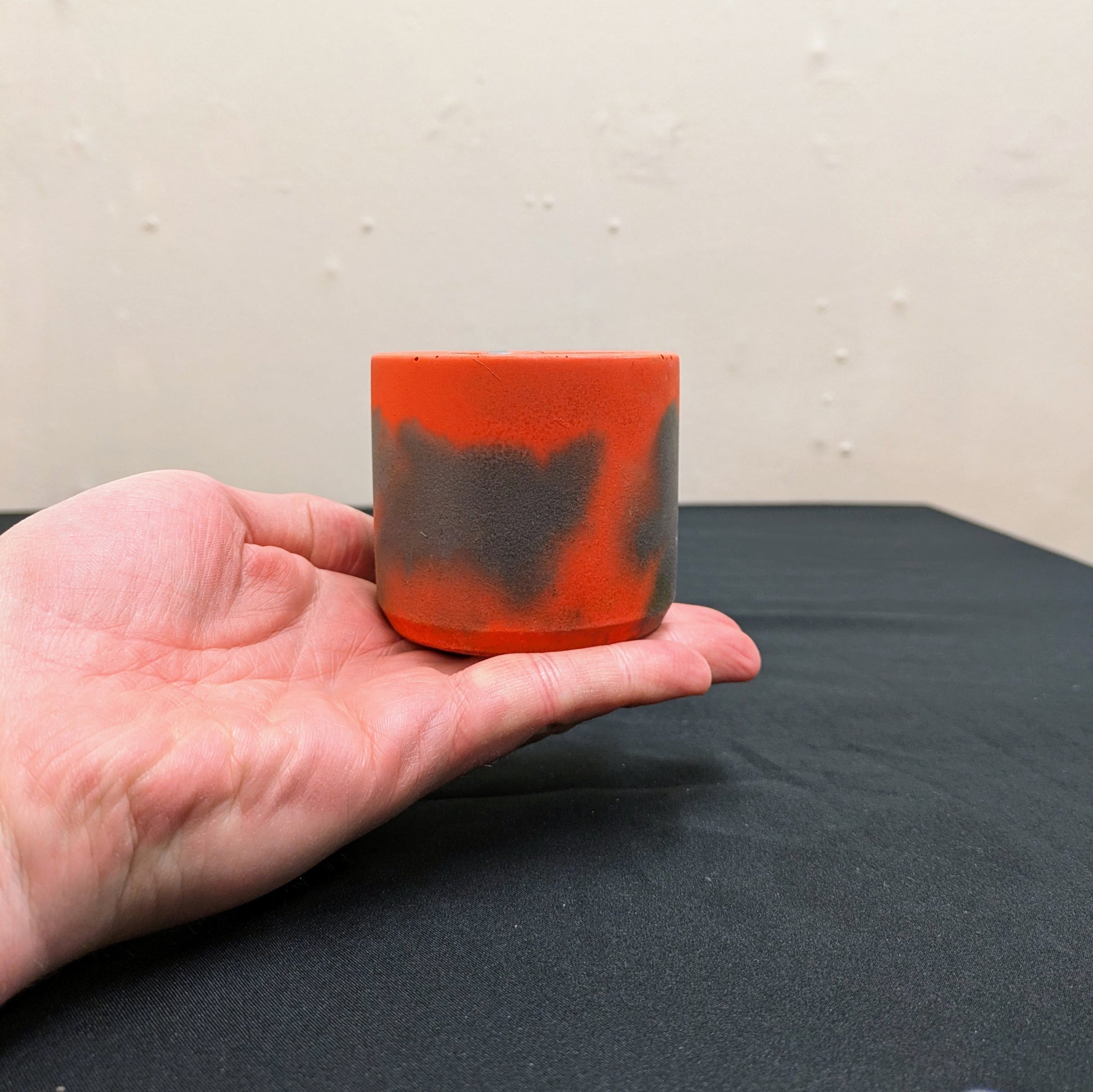 The picture shows a grey and orange marbled concrete plant pot, it is a small pot being held at the end of a hand to show scale.