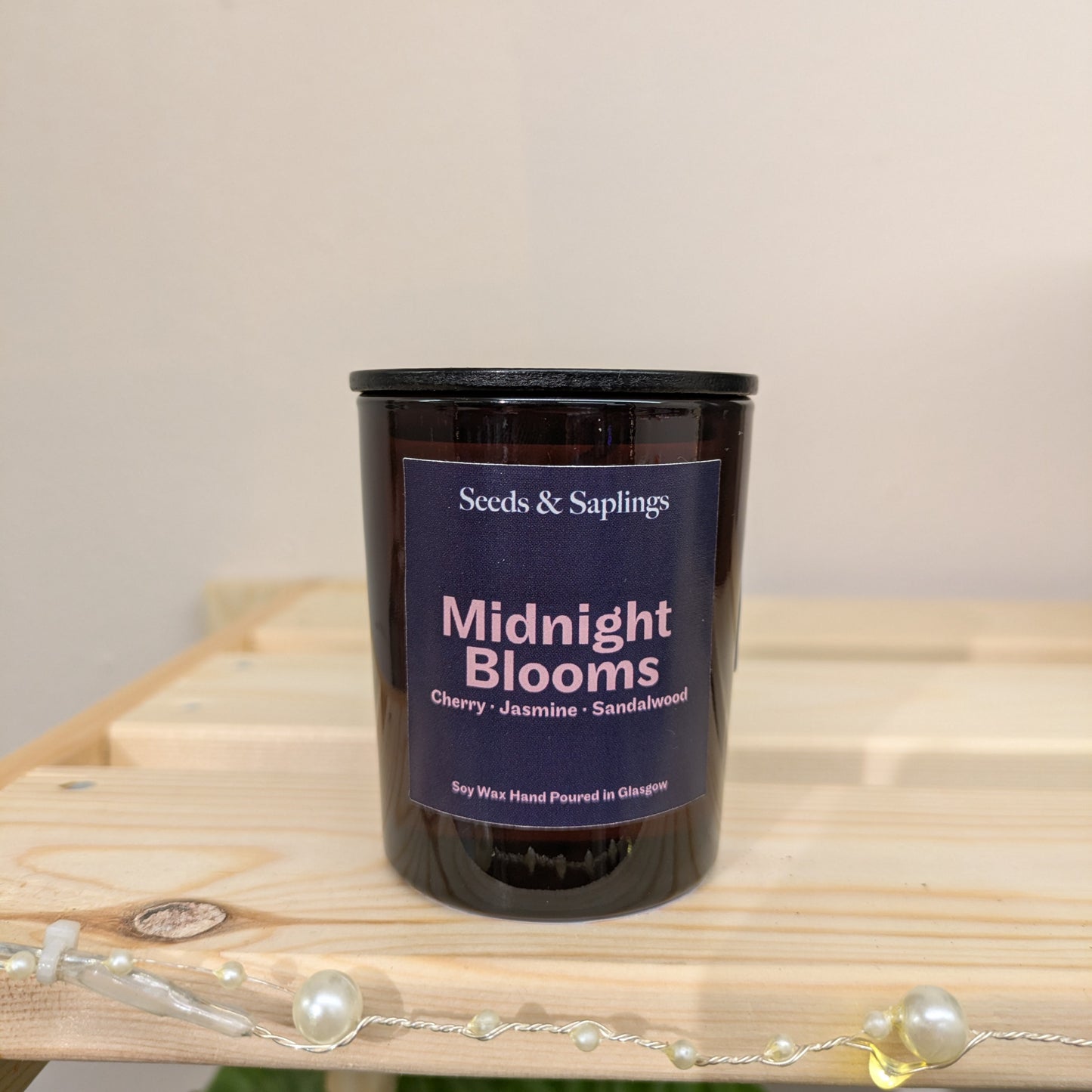 The image shows an amber candle jar with a black lid on a wooden shelf. The label says ‘Midnight Blooms’, ‘Cherry, Jasmine, Sandalwood’ and ‘Soy Wax Hand Poured in Glasgow’.
