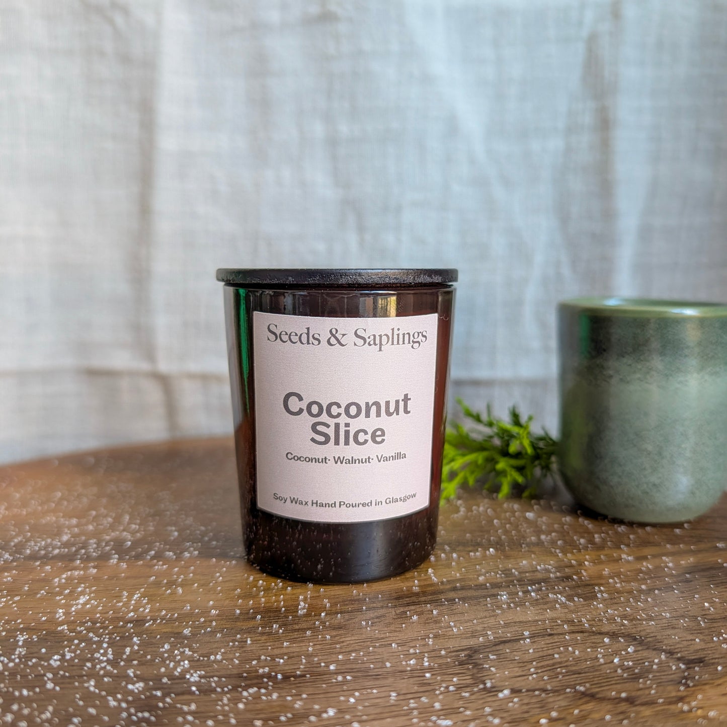 Candle labeled 'Coconut Slice' by 'Seeds & Saplings' on a wooden surface with a neutral background.