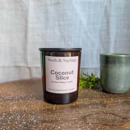 Candle labeled 'Coconut Slice' by 'Seeds & Saplings' on a wooden surface with a neutral background.